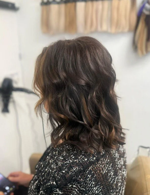 Side view of a woman with shoulder-length wavy brown hair with highlights, sitting in a hair salon.