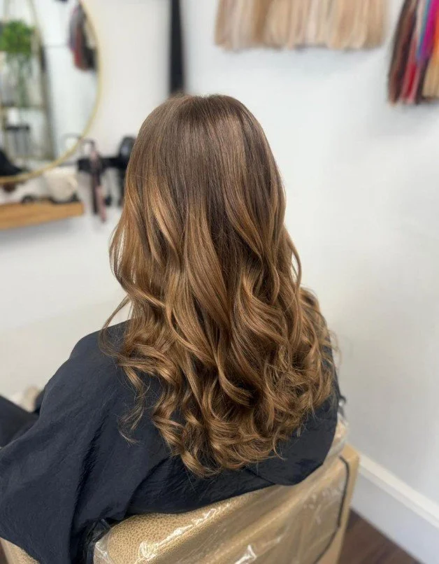 Back of a woman with long, wavy, light brown hair sitting in a salon chair with a black cape.