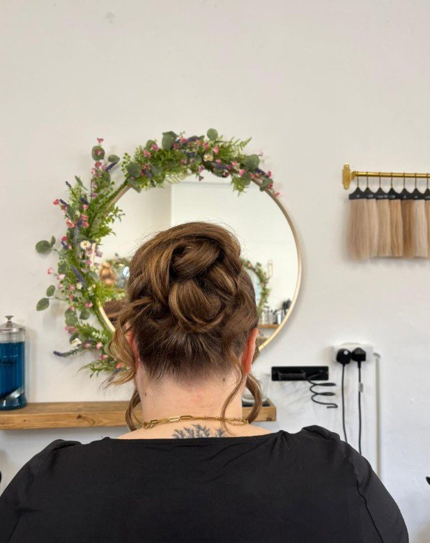 Back of a woman with an intricate braided and curled updo hairstyle, sitting in front of a round mirror decorated with a floral wreath, in a hair salon.