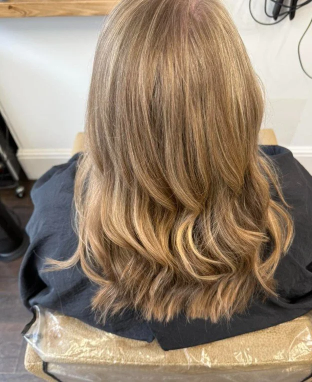 Back view of a woman with long, wavy, blonde hair sitting in a salon chair, wearing a black cape.