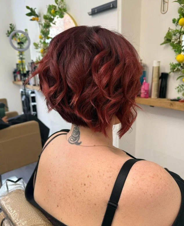 A woman with short, wavy, auburn hair sitting in a hair salon with a tattoo on her neck, wearing a black top with thin straps.