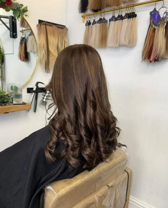 Back view of a woman with shoulder-length, wavy brown hair sitting in a salon chair.