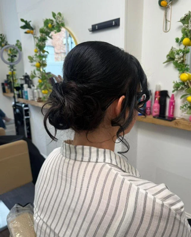 A woman with dark hair styled in a loose bun, facing away in a salon with a wall shelf holding hair products, decorative plants, and a mirror.