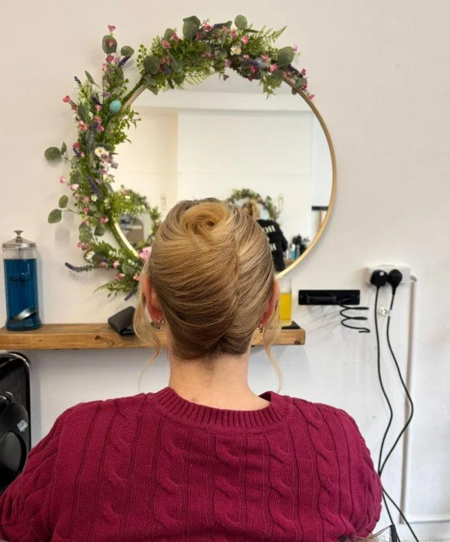 Woman with blonde hair styled in an elegant updo, wearing a red cable-knit sweater, sitting in front of a circular mirror decorated with pink and purple flowers and greenery in a salon or beauty shop.