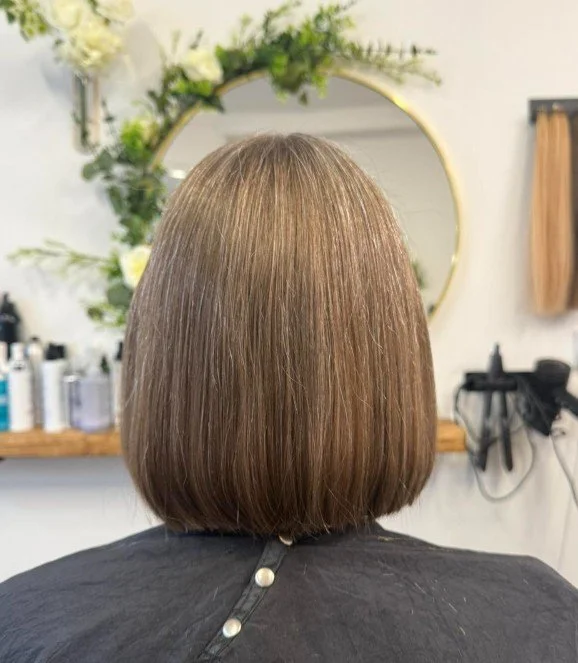 Back of a woman with straight, shoulder-length brown hair in a salon, with a round mirror and salon products in the background.