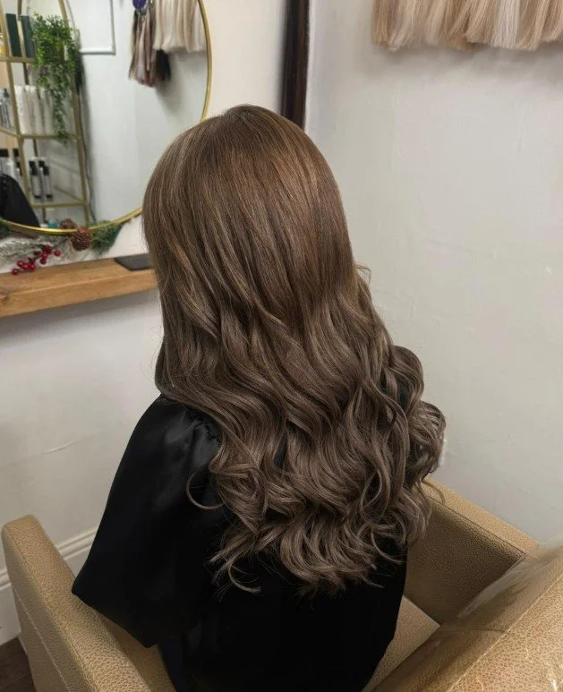 Back view of a woman with long, wavy, light brown hair sitting in a salon chair
