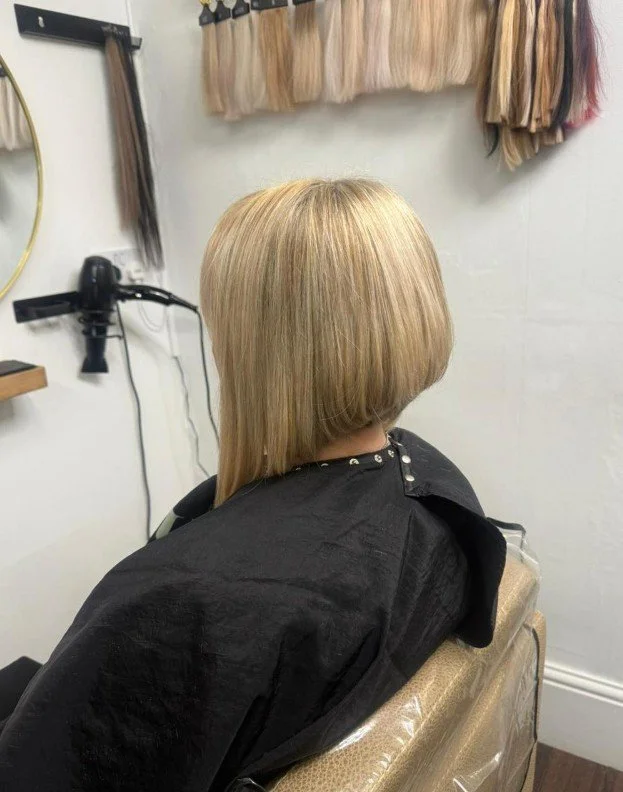 Woman with a freshly cut blonde bob hairstyle sitting in a salon chair, facing away from the camera, with hair color samples hanging on the wall behind her.