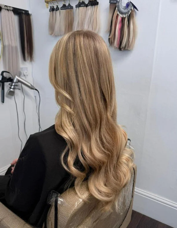 A woman with long, wavy blonde hair sitting in a salon chair. Hair extensions or wigs are hanging on the wall behind her.