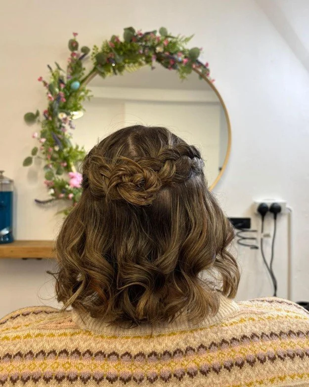 Back view of a woman with wavy brown hair in a half-up braided hairstyle, sitting in front of a round mirror decorated with pink and purple flowers.