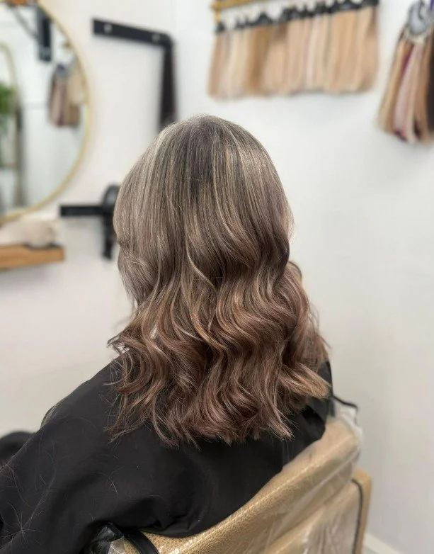 Back view of a woman with shoulder-length wavy hair in a salon, with color-treated hair featuring subtle highlights, and a salon mirror and hair swatches in the background.
