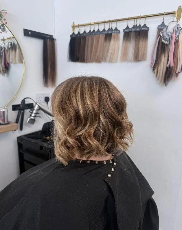 A woman with short, wavy light brown hair sitting in a salon chair, facing away. Hair color samples on the wall behind her.