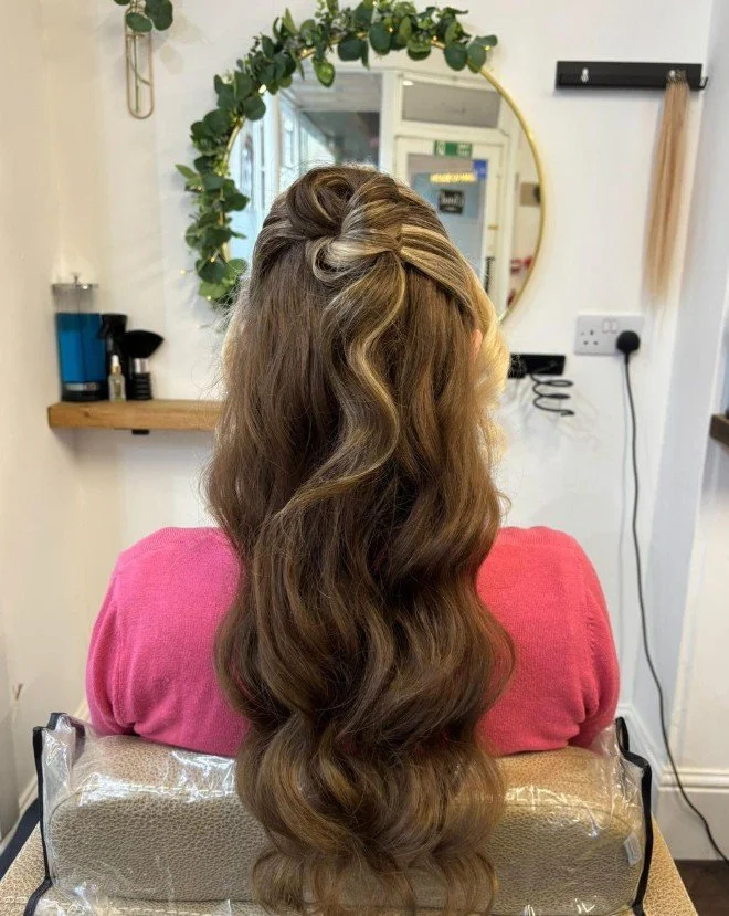 A woman with long, wavy brown hair sitting in a salon chair, facing away from the mirror. Her hair features a half-up, twisted hairstyle at the crown. The salon decor includes a round mirror surrounded by green foliage, a shelf with hair products, an