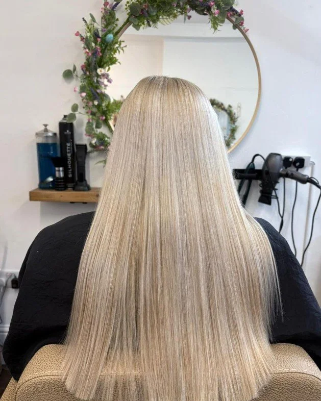 Back view of a person with long, straight, blonde hair in a salon, with a circular mirror decorated with flowers, and hairstyling tools on a shelf and wall.