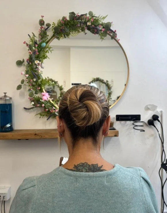 A woman with blonde hair styled in a bun, sitting in front of a round mirror decorated with pink and purple flowers, in a salon or similar setting.
