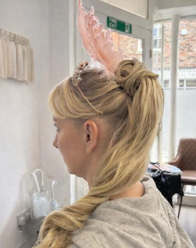 A woman with elaborate blonde hair styled in a half-up, half-down fashion, wearing a large pink feathered headband and decorative hair accessories.