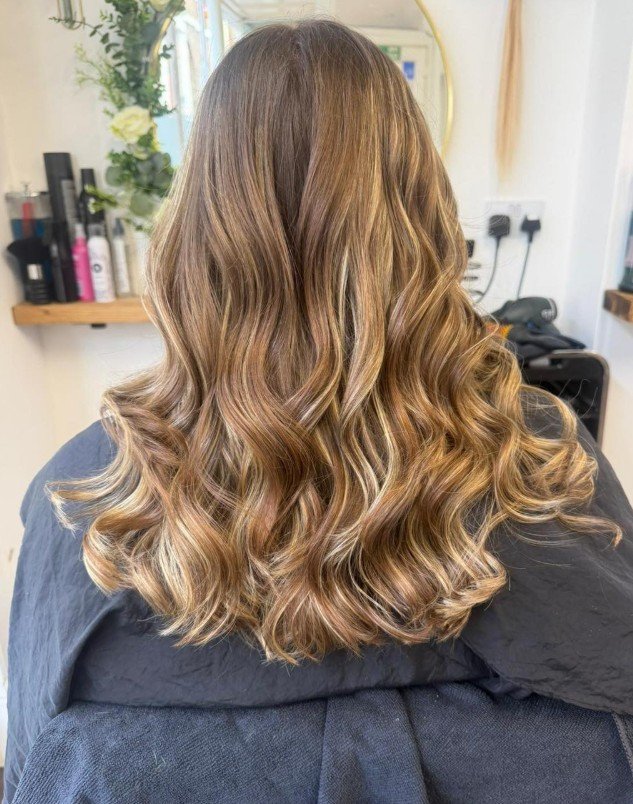Back of a woman's head showing styled wavy, caramel-colored hair with highlights in a salon setting.