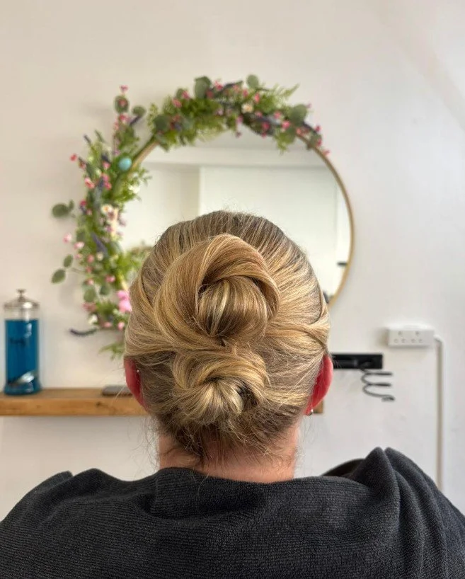 Woman with blonde hair styled in a bun, sitting in front of a round mirror decorated with pink and green floral wreath.