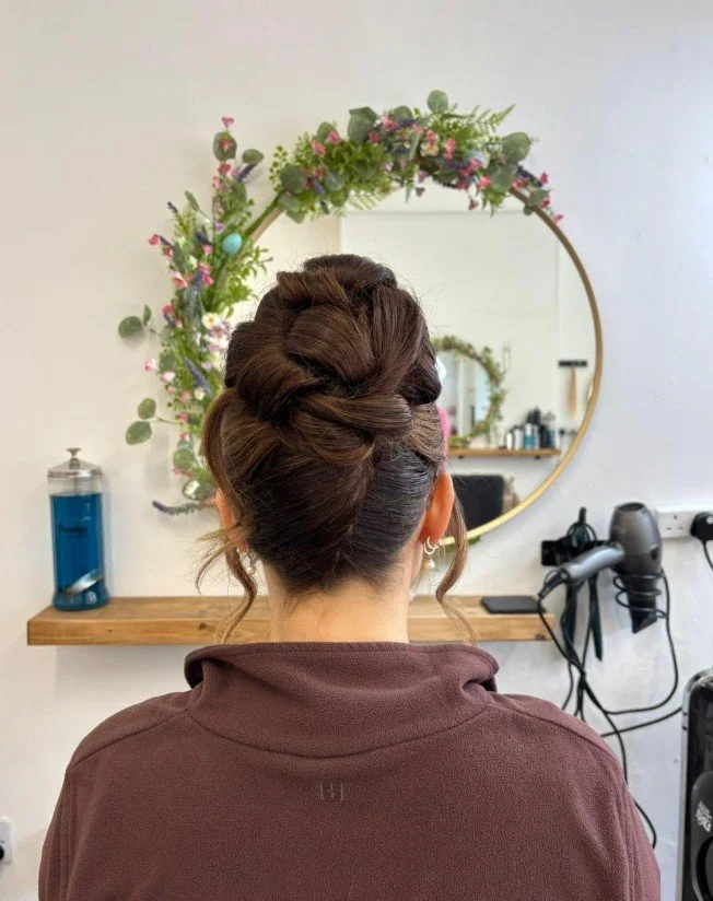 Back of a woman with an intricate braided updo hairstyle, sitting in front of a round mirror decorated with floral arrangements, in a room with salon equipment.