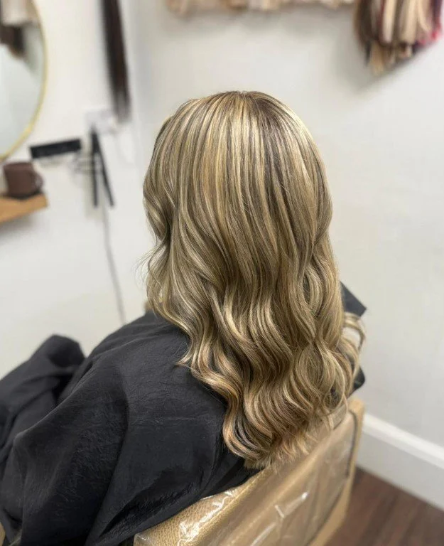 A woman with blonde, wavy hair sitting in a salon chair, wearing a black cape, in a salon with mirrors and hair tools visible in the background.