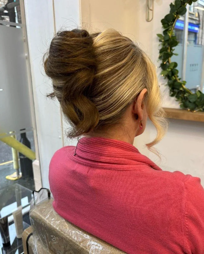 Back view of a woman with blonde hair styled in an elegant updo, wearing a pink sweater and small earrings, sitting in a salon or similar setting with a mirror and plant visible in the background.