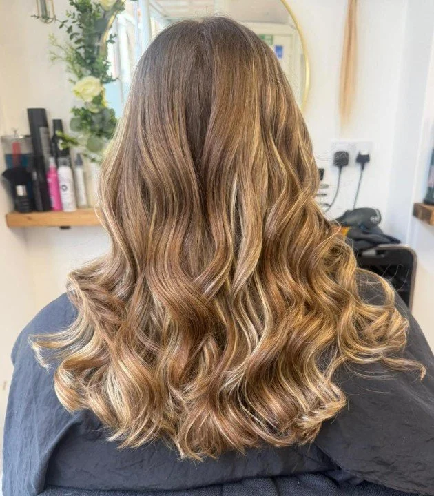 Back view of a woman with long, wavy, ombre hair transitioning from light brown to blond at the ends, in a salon setting.