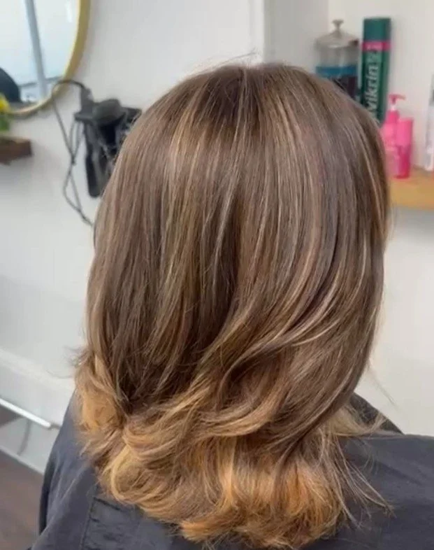 Back view of a woman with shoulder-length, wavy, light brown hair in a salon.