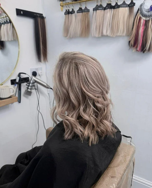 Woman with wavy blonde hair sitting in a salon chair, preparing for a hair styling or coloring session, with hair swatches hanging on the wall behind her.