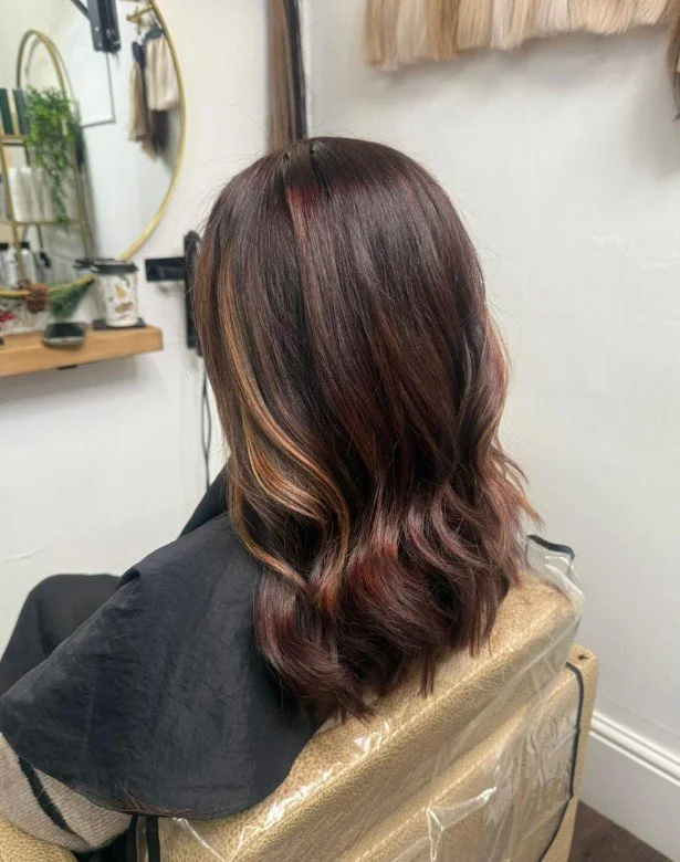 Woman with shoulder-length, wavy hair dyed in shades of dark brown with caramel highlights, seated in a salon chair.