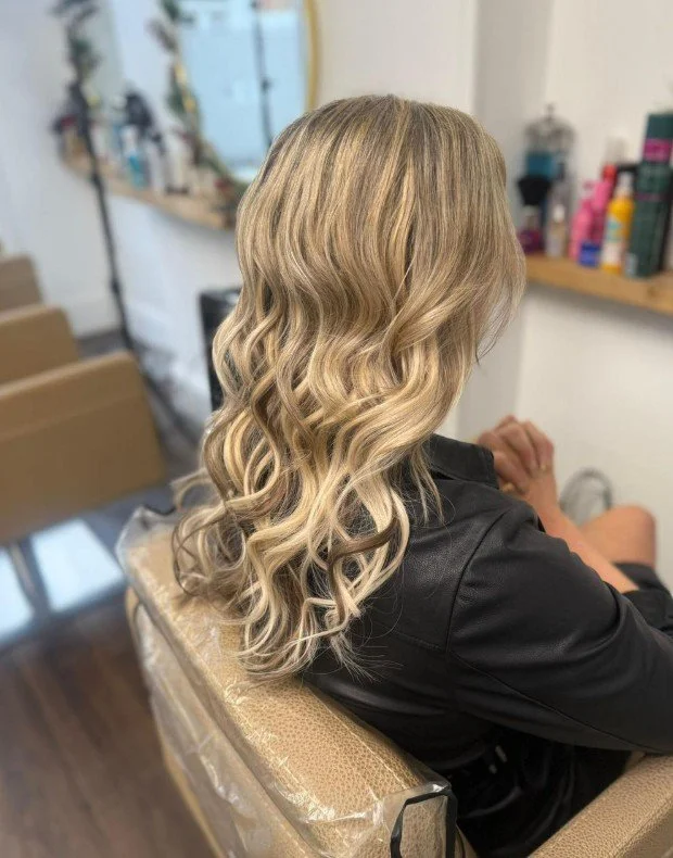 Back view of a woman with long, blonde, wavy hair sitting in a hair salon.