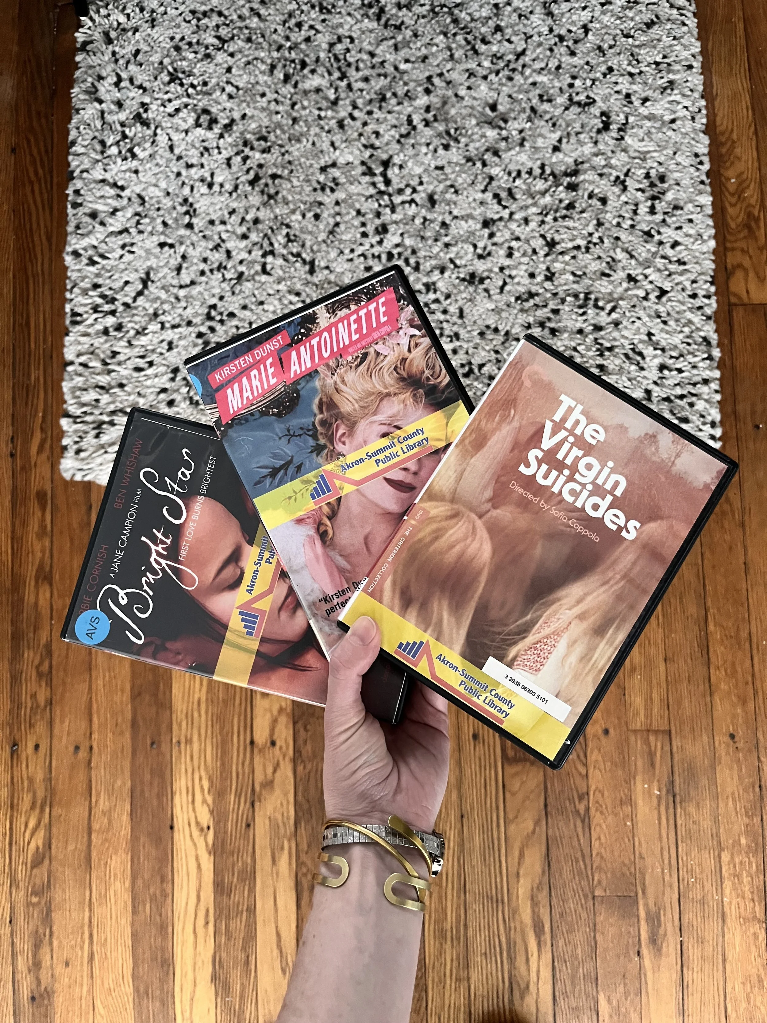A hand holding three DVD cases—"Marie Antoinette," "Bright Star," and "The Virgin Suicides"—above a textured rug and wooden floor.  Transcribed Text:      Marie Antoinette     Bright Star     The Virgin Suicides     Akron-Summit County Public Library