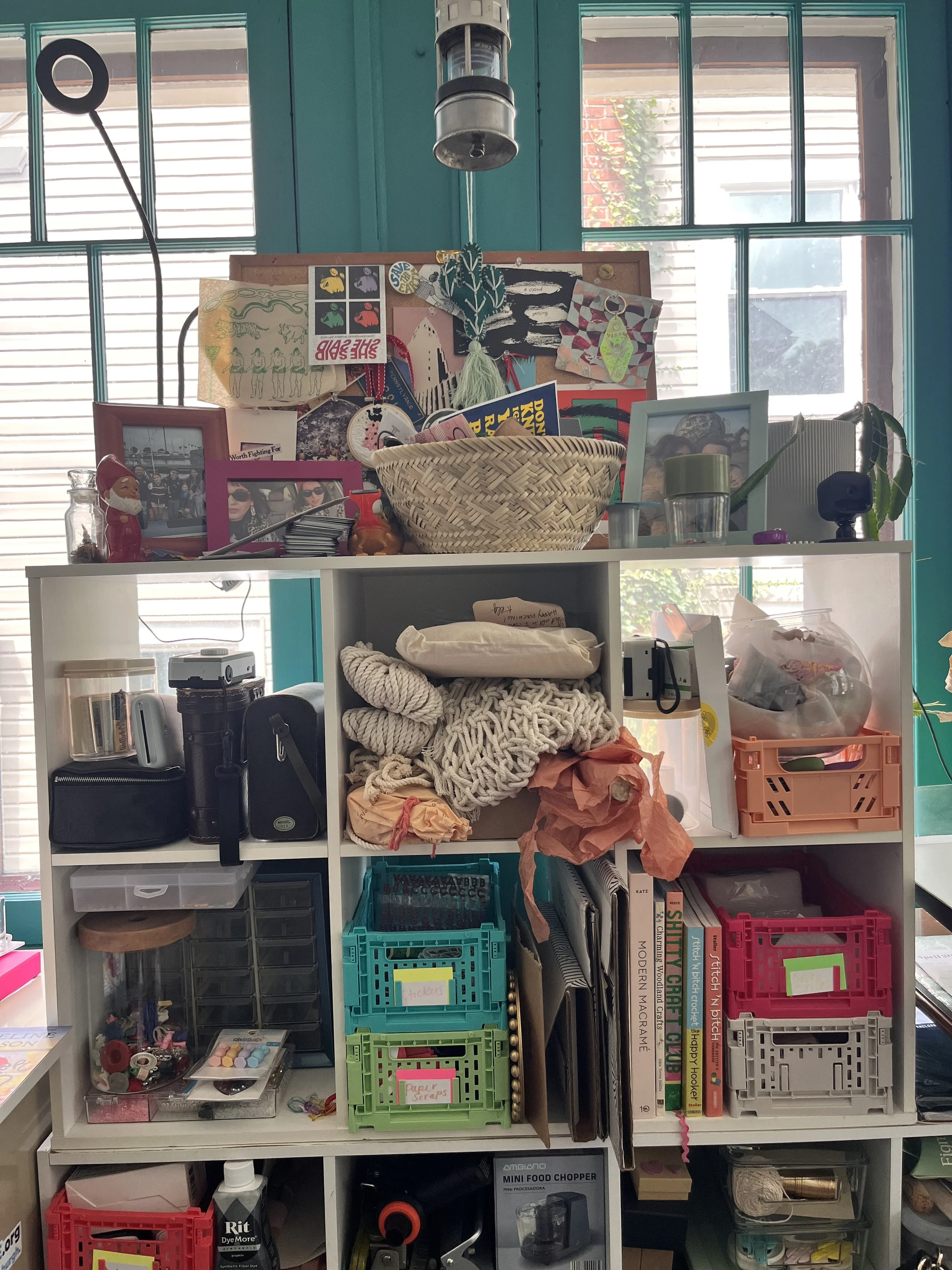 A cluttered shelving unit with a variety of colorful containers, craft supplies, and decorative items against teal walls and large windows.