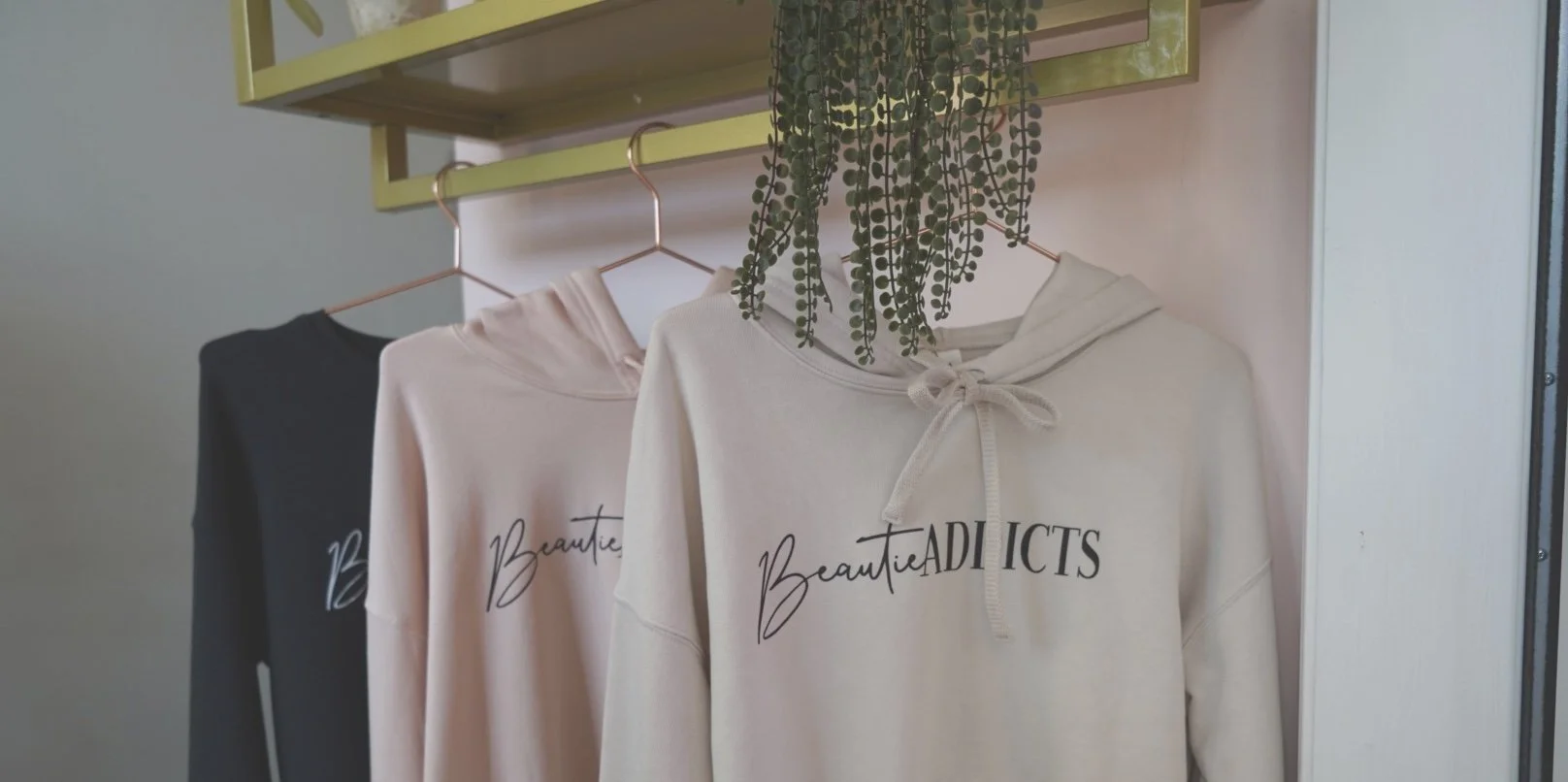 Three hoodies hanging on a rack, each with the words 'BeautieADDICTS' printed on them, with a green plant hanging above.