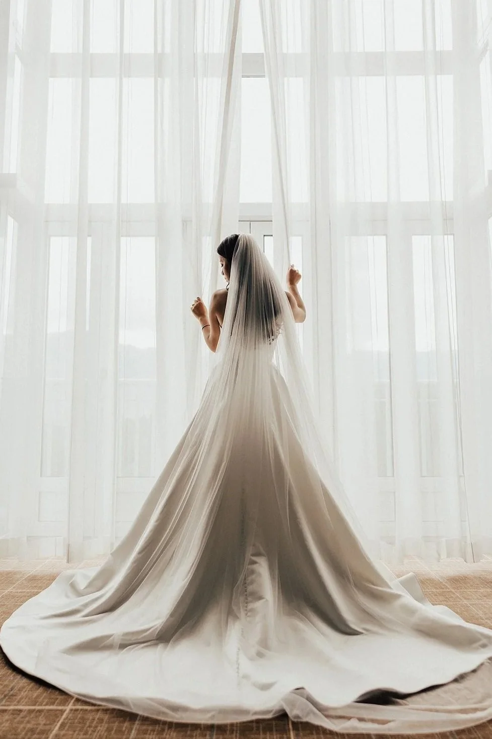 A Sea to Sky bride in a white wedding gown and veil standing in front of large window with sheer white curtains, backlit by natural light.