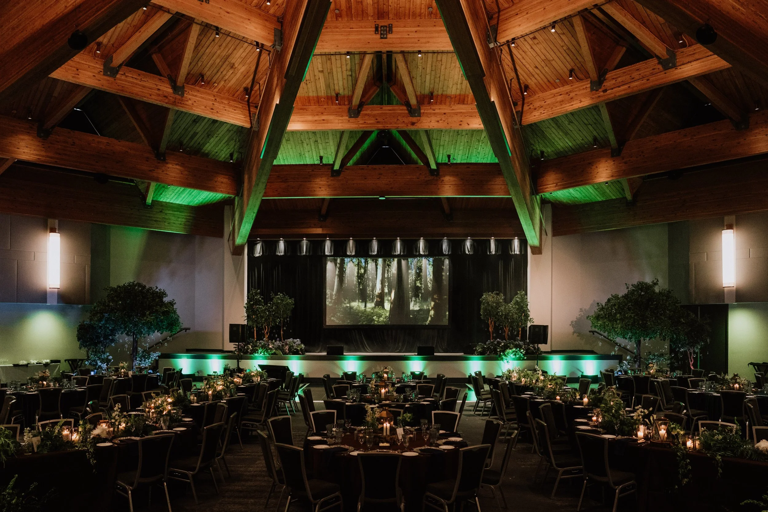 An indoor event hall with a high wooden, vaulted ceiling illuminated with green lighting. The room features a stage with a large screen displaying a forest scene, and tables decorated with floral centerpieces and candles, set for a formal event.
