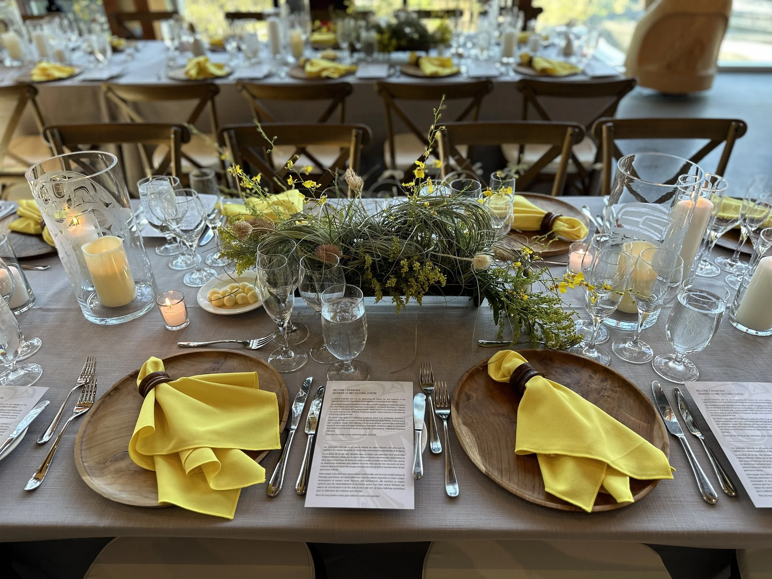 A formal banquet table set with yellow napkins, clear glassware, candles, and a floral centerpiece with yellow and green flowers. Wooden chargers are at each place setting with printed menus and silverware.