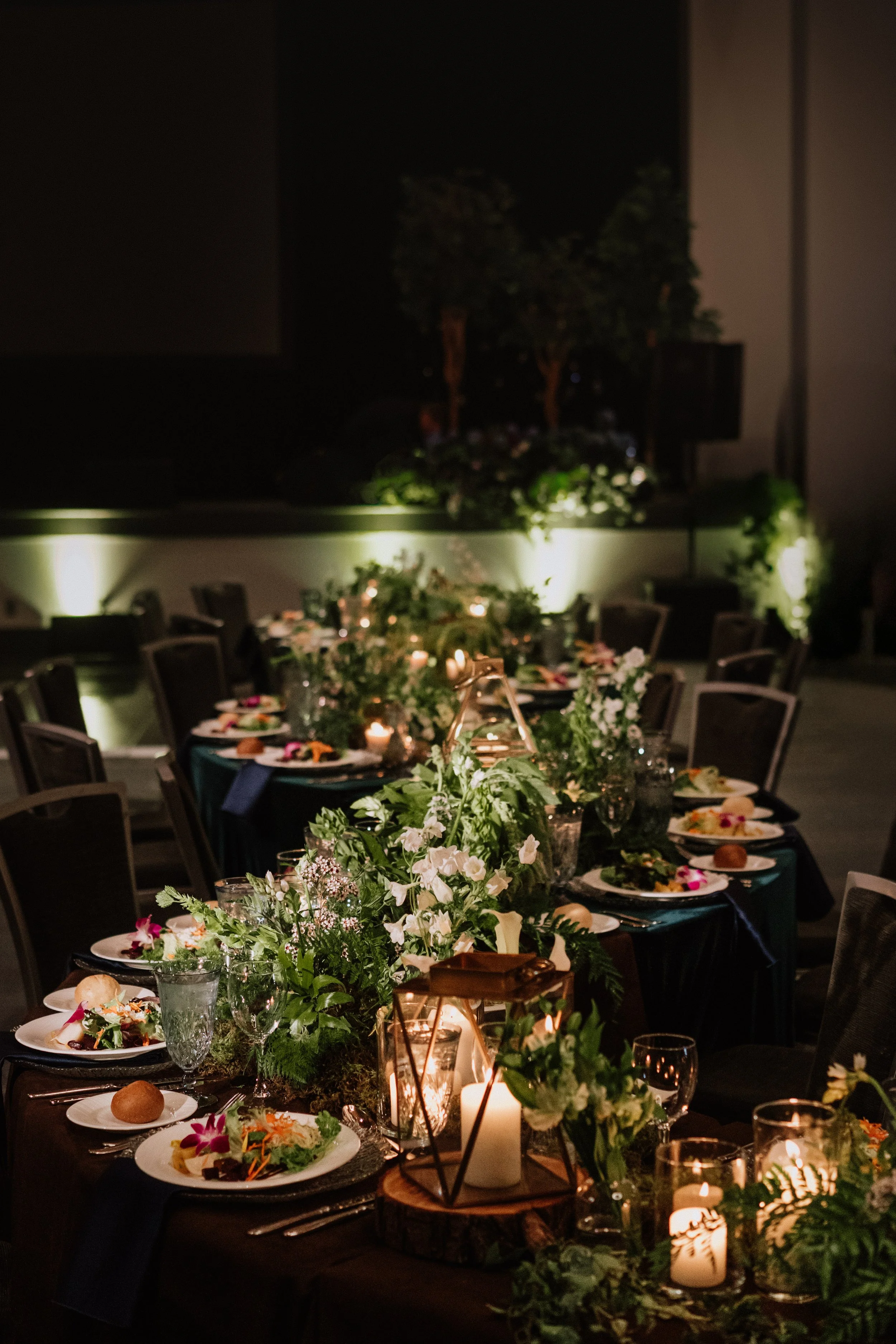 A beautifully decorated indoor dining table set for an event, with candles, floral centerpieces, and place settings, in a dimly lit environment.