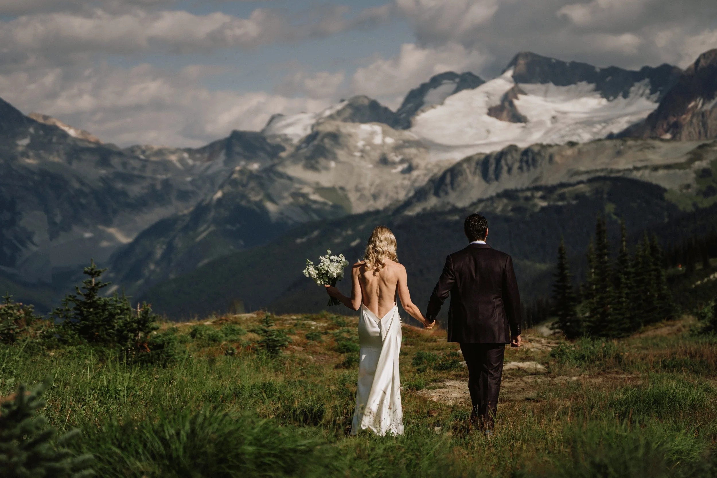 A bride and groom holding hands and walking in a grassy field with Whistler mountains in the background.