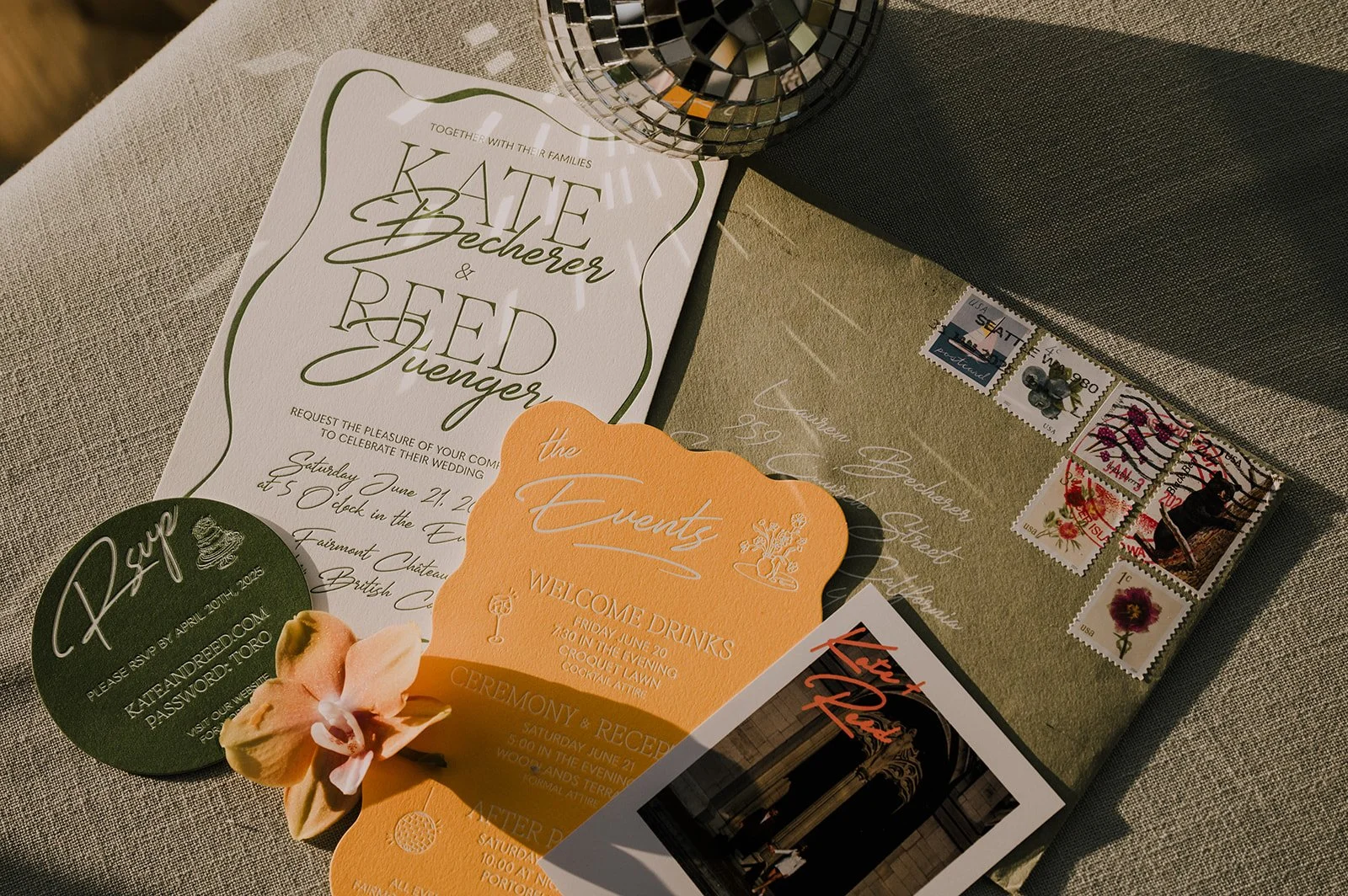 Wedding invitation and related stationery including an envelope with stamps, a green circular card, and an orange scalloped card, laid on a table with a metallic candle holder.
