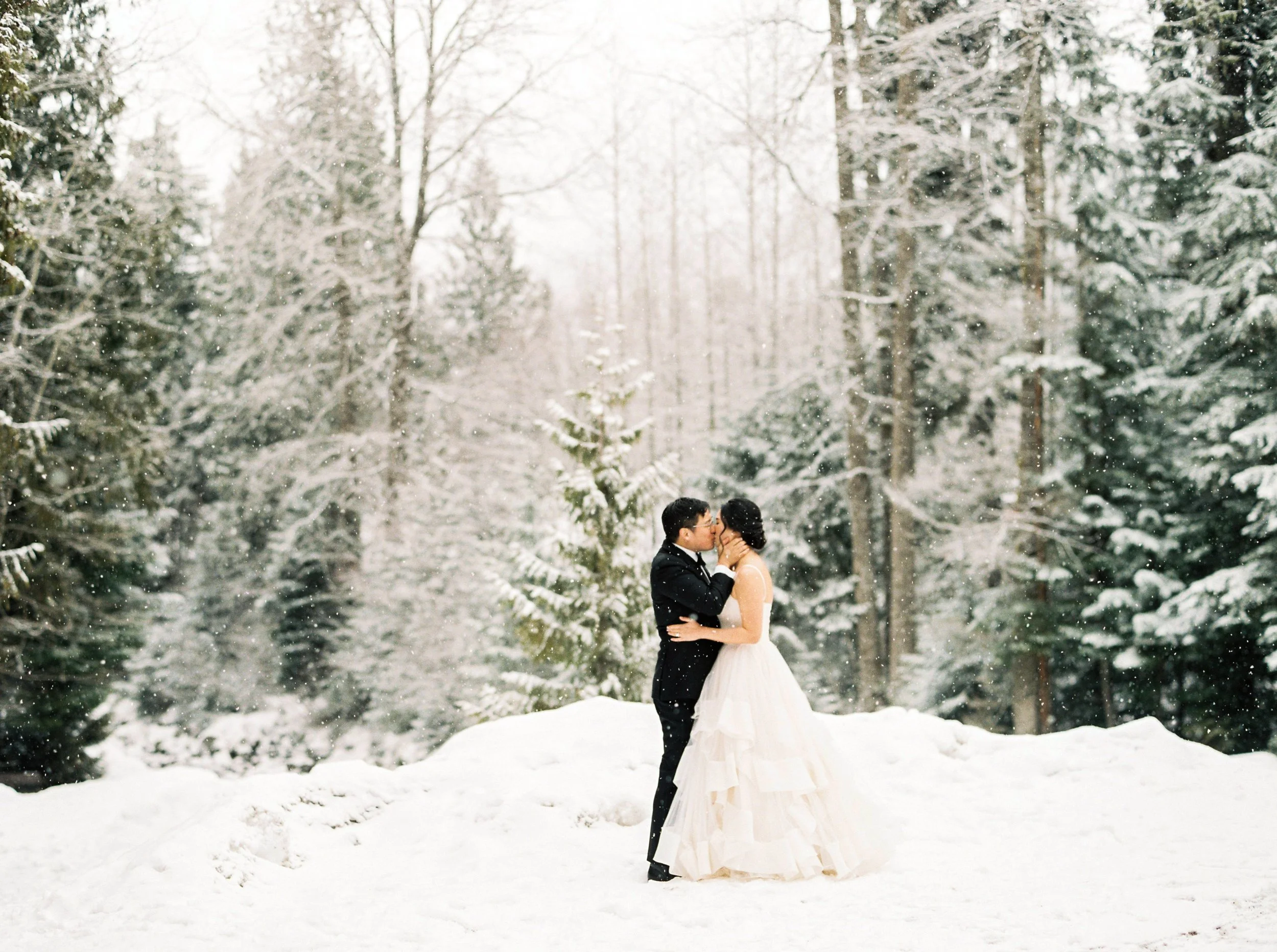 A bride and groom kiss in a snow-covered forest during winter.