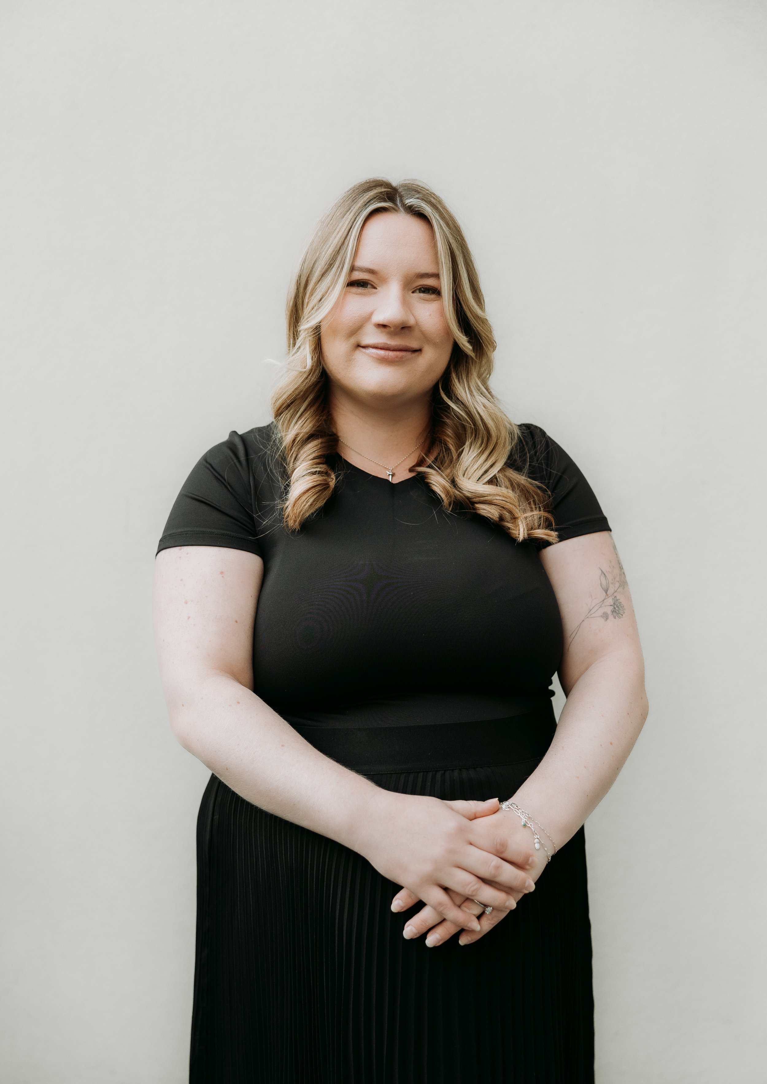 A woman with blonde hair styled in loose curls, wearing a black top and black pleated skirt, standing against a plain white wall with her hands clasped in front of her, smiling softly.