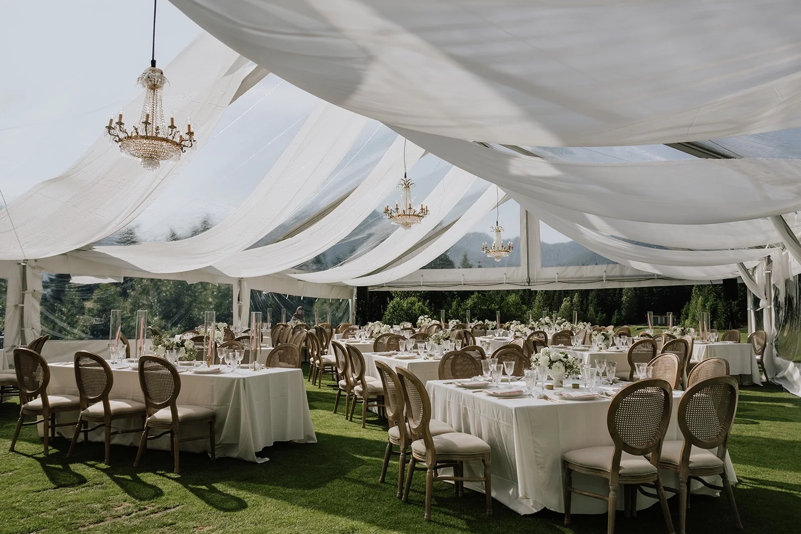 Elegant outdoor event setup under a large white tent with chandeliers, round and rectangular tables with white tablecloths, floral centerpieces, and cushioned chairs, set on a grassy area with trees in the background Whistler.
