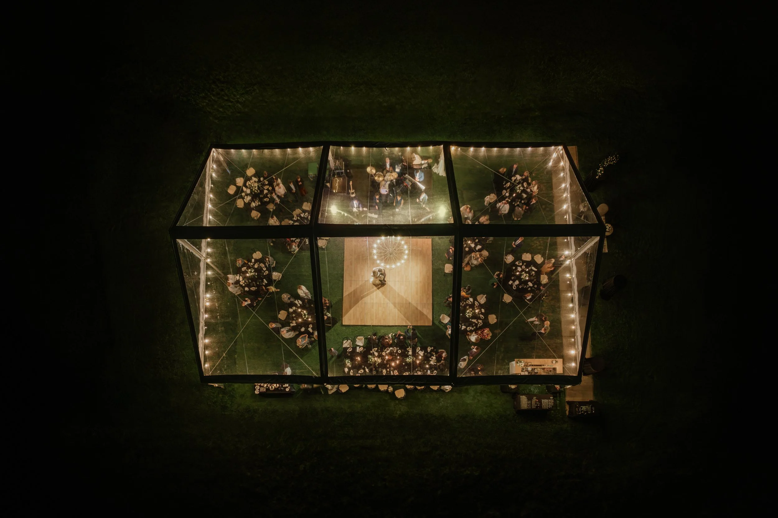 An aerial view of a glass enclosure at night, with people inside attending a party or gathering, illuminated by string lights.