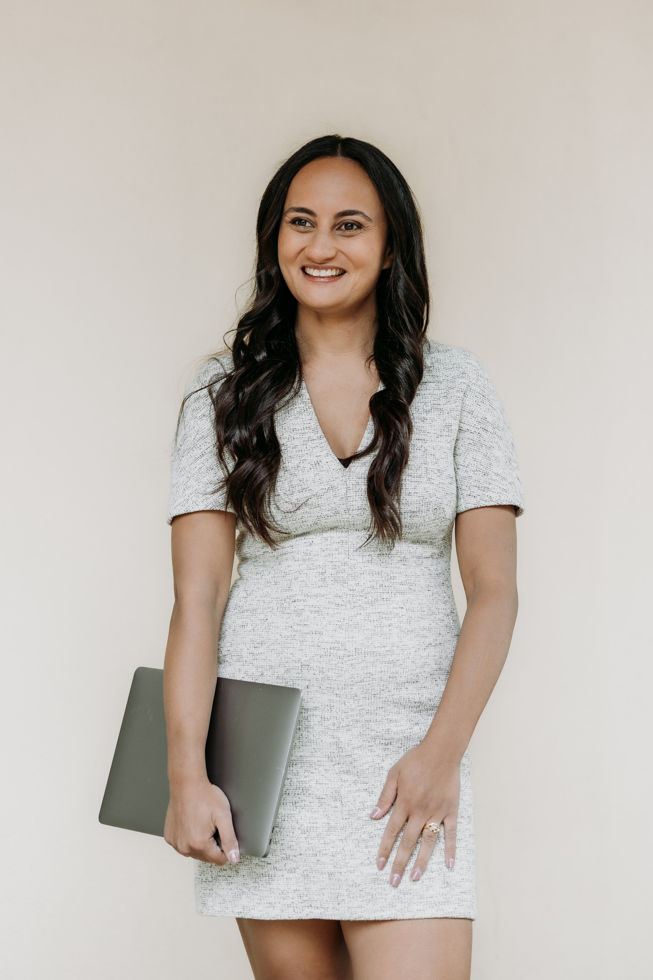 A woman with long dark hair, wearing a light-colored dress, holding a closed laptop, smiling, standing against a plain background.