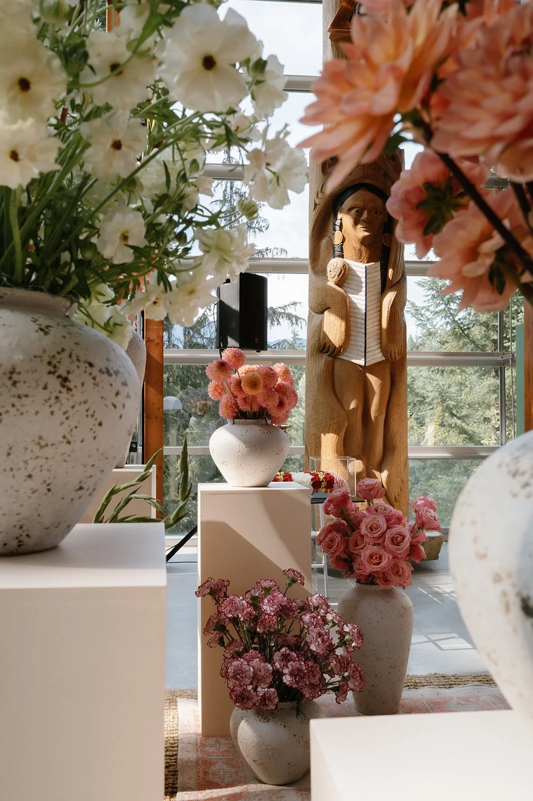 Vases with white, pink, and red florals arranged in a Whsitler gallery with large windows, a wooden sculpture of a human figure, and trees visible outside.