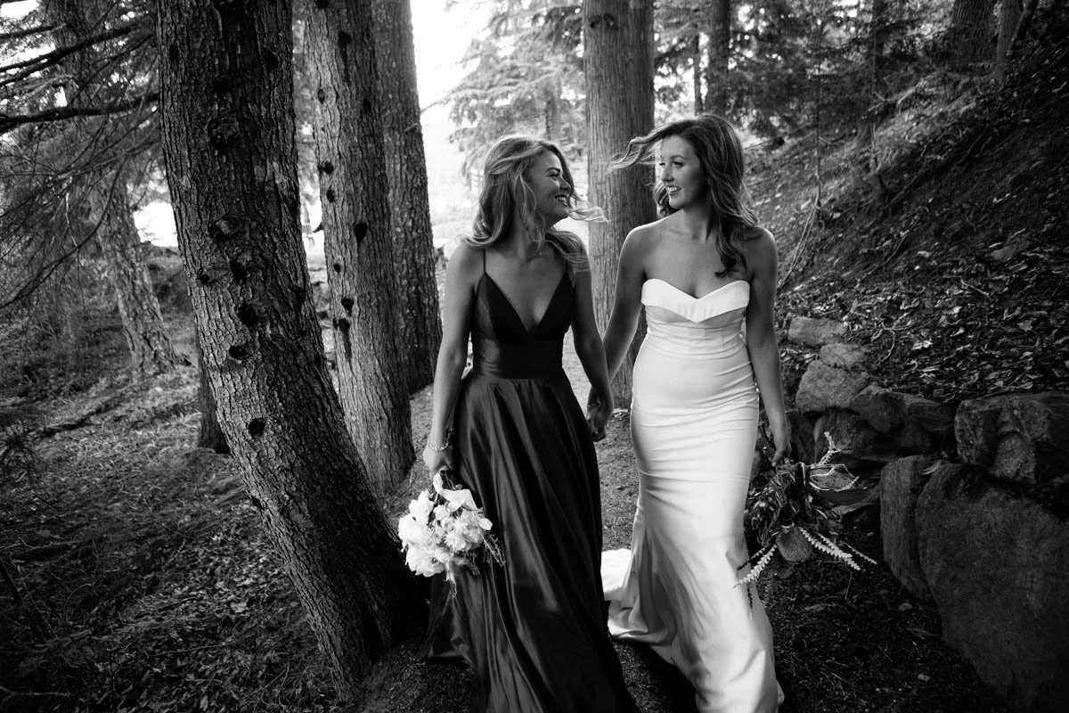 Two women in wedding dresses walking through a wooded area, smiling and holding hands, one in a dark gown and the other in a white gown, with trees and rocks in the background.