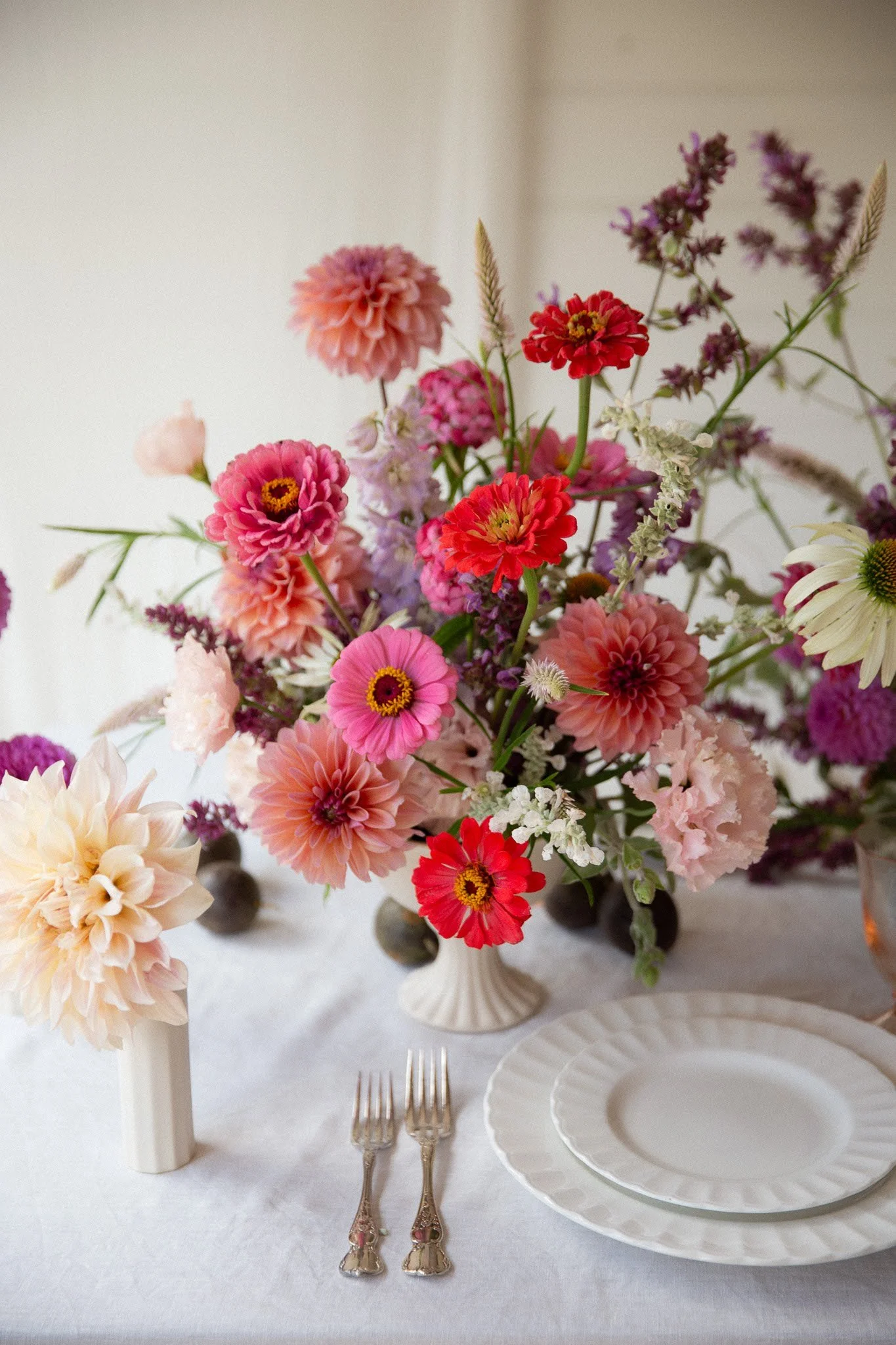 Vase with pink, red, purple, and white flowers on a table with white plates, silver forks, and a small white vase with large cream-colored flowers.