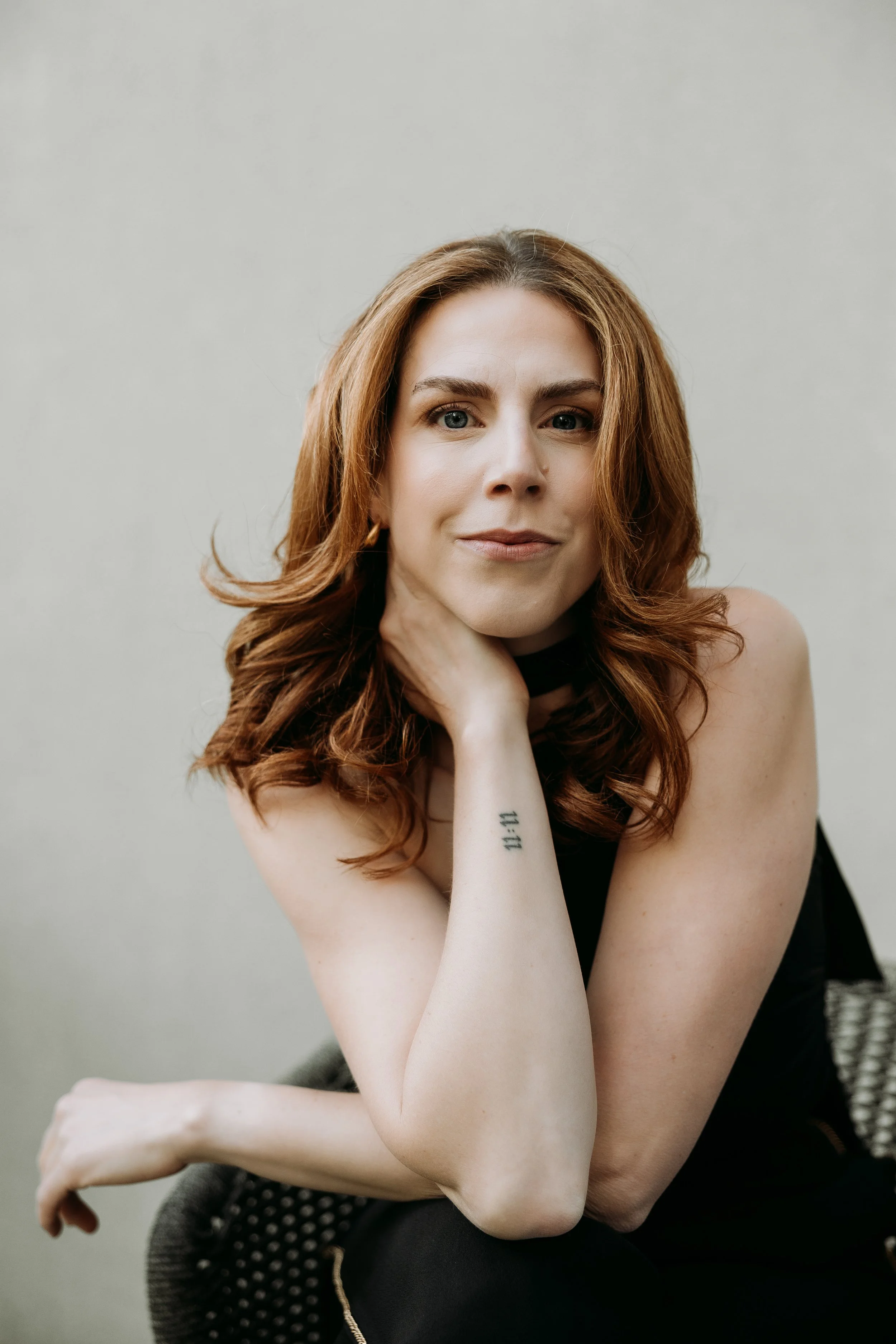 Portrait of a woman with wavy red hair, sitting with her chin resting on her hand, wearing a black top and gold earrings, against a plain light background.
