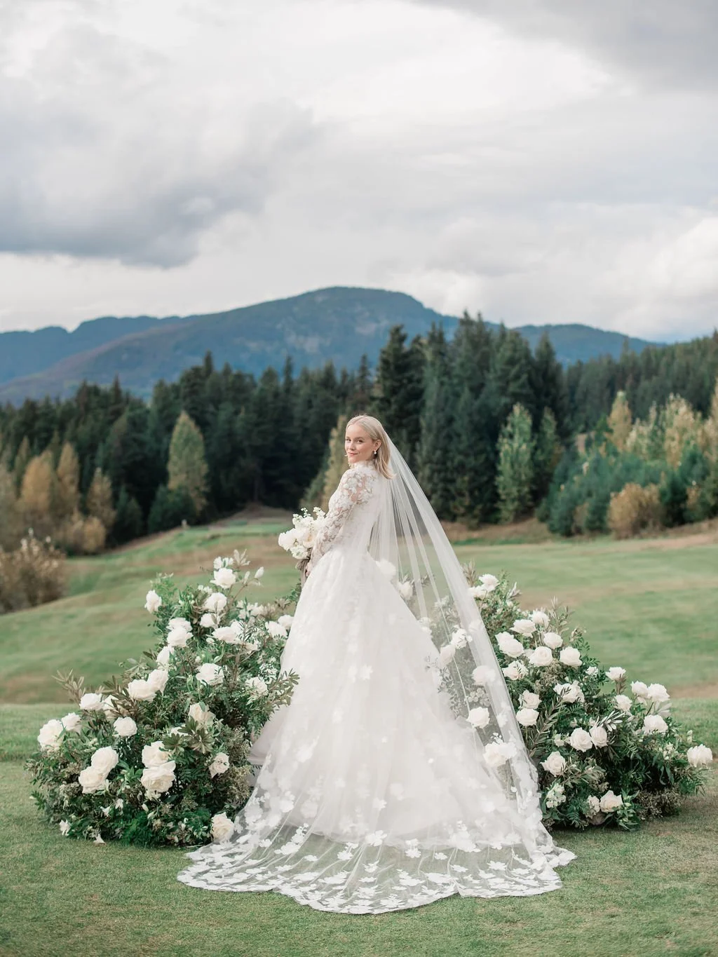 A bride in a white wedding dress with lace sleeves and a long veil stands outdoors on a grassy field, surrounded by white flowers, with a backdrop of green trees and Whistler mountains under cloudy sky.
