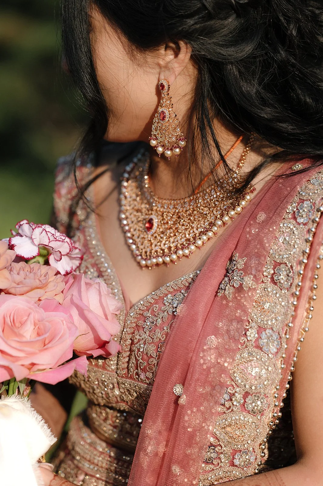 A woman dressed in traditional bridal attire with intricate jewelry and a pink embroidered saree, holding a bouquet of pink and white flowers.