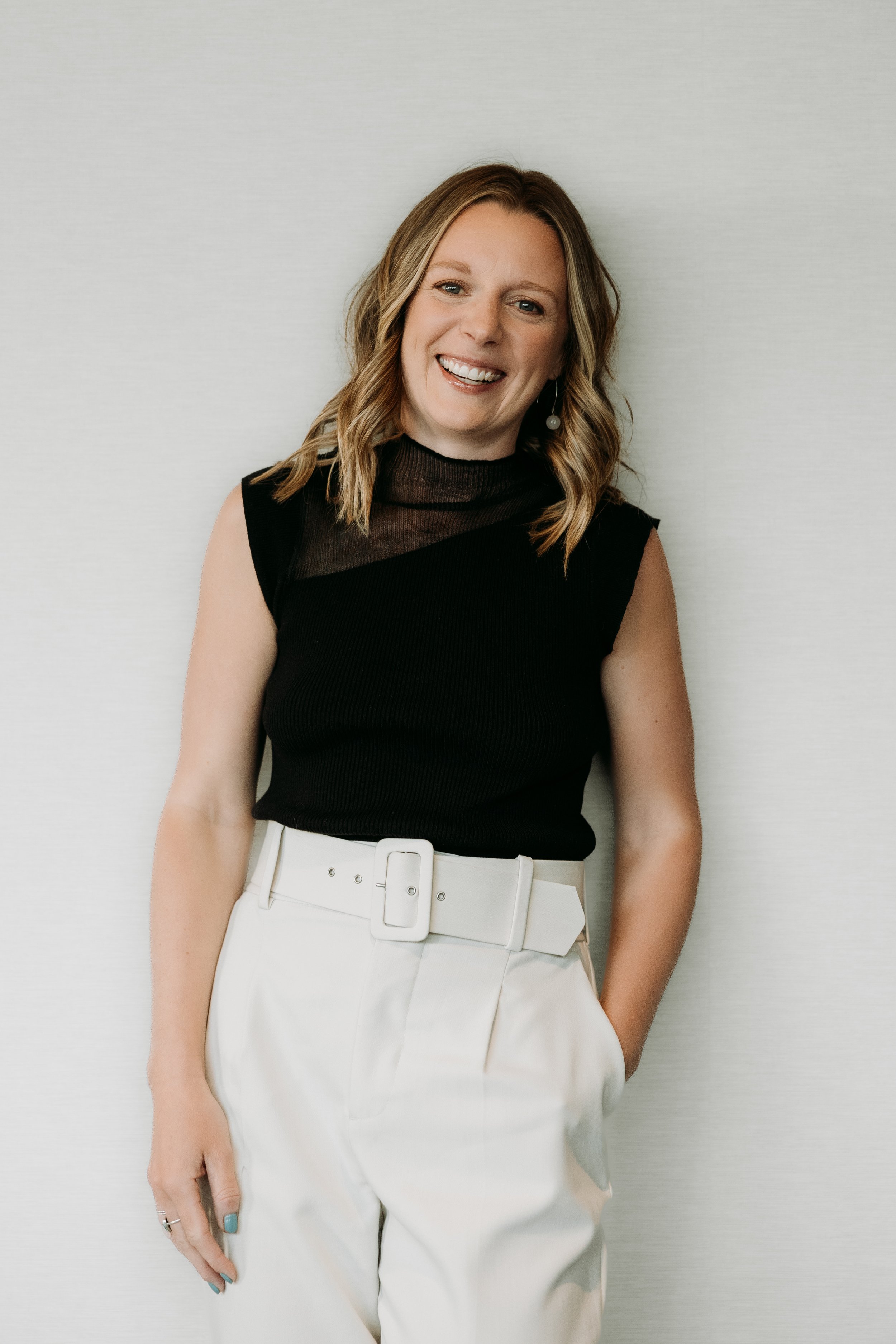 A woman with wavy, shoulder-length hair wearing a sleeveless black top with a sheer high neckline and white high-waisted pants with a large belt, smiling at the camera.